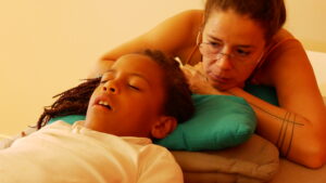 In this photo, a moment of care in biodynamic craniosacral therapy is captured.
A child is lying on their back, with their head comfortably resting on a blue cushion. Their features are relaxed, and they appear deeply at ease, illustrating a state of release often sought during a craniosacral therapy session.
Beside the child, the therapist offers an attentive and caring gaze. Leaning on her arm, she embodies a calm and reassuring presence. Her hands are not visible in the image, but her posture suggests the subtle listening and supportive approach that characterize this gentle therapy.
The simple and warm environment highlights the sense of serenity and safety experienced during craniosacral care, where attention is given to the body’s natural rhythm and its capacity for self-regulation.
This image reflects the ability of craniosacral therapy to create a space for relaxation and harmony for everyone, including children.